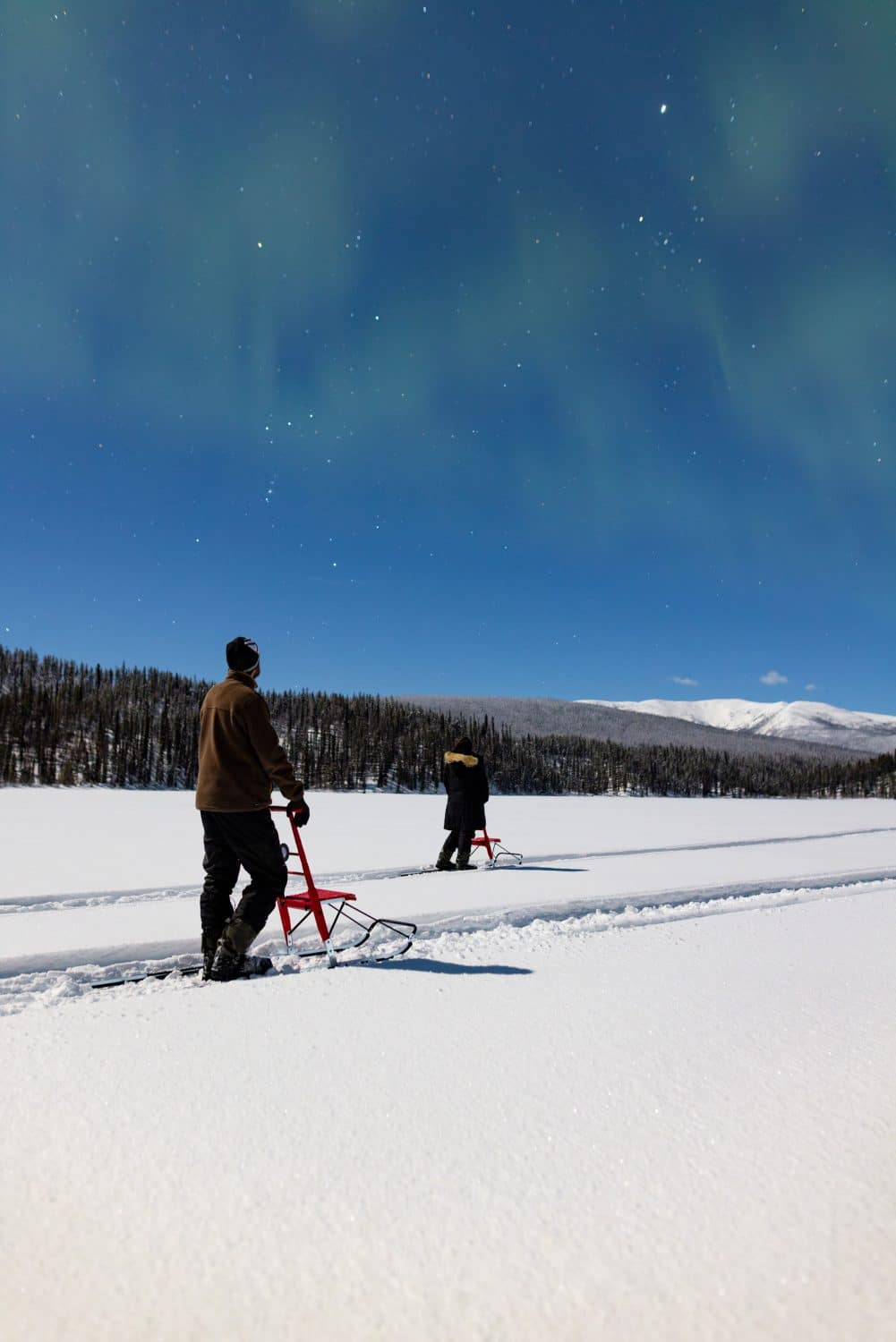 Two people ice fishing under auroras